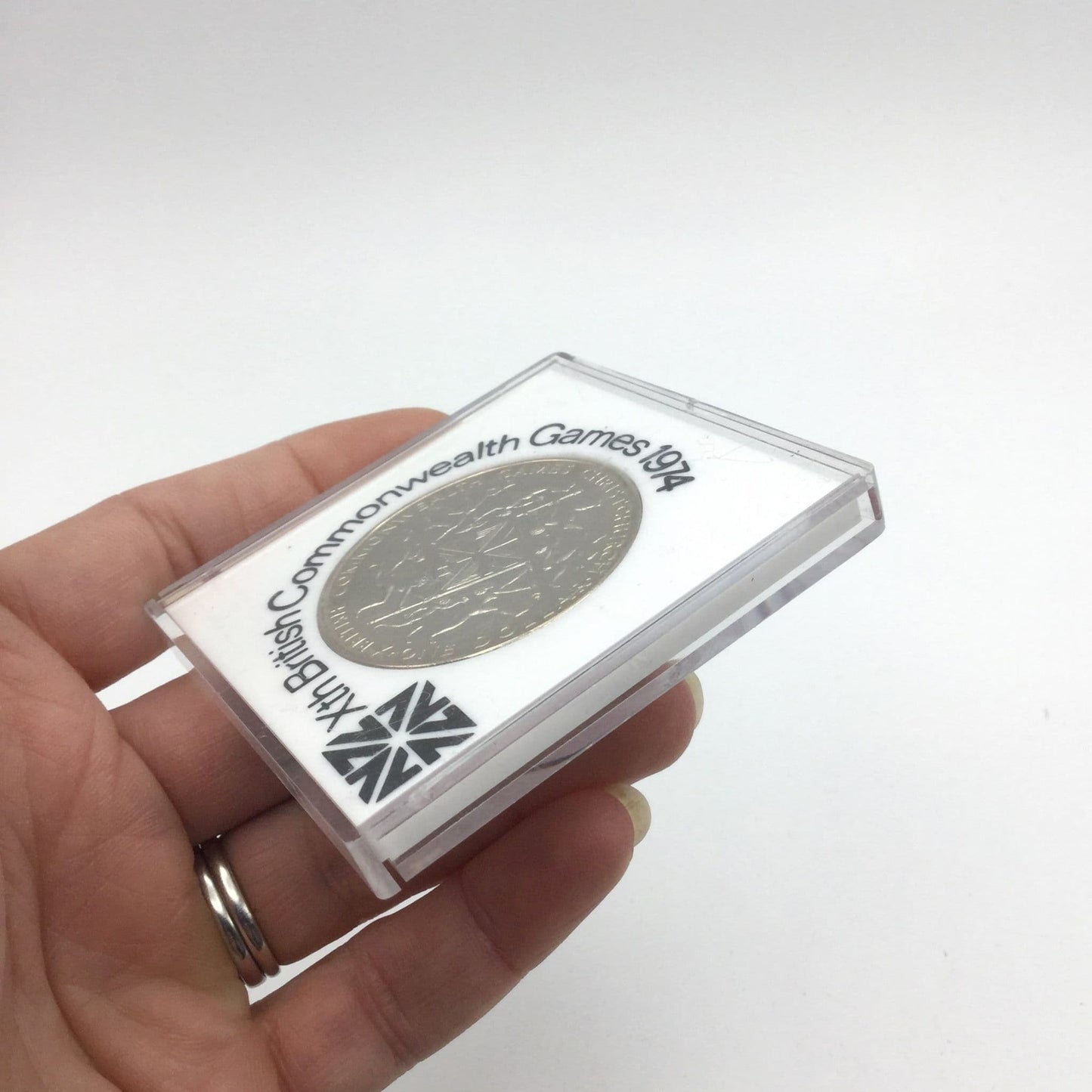 Side view of One dollar coin in a white case with 10th British Commonwealth Games 1974 around it held in a hand.