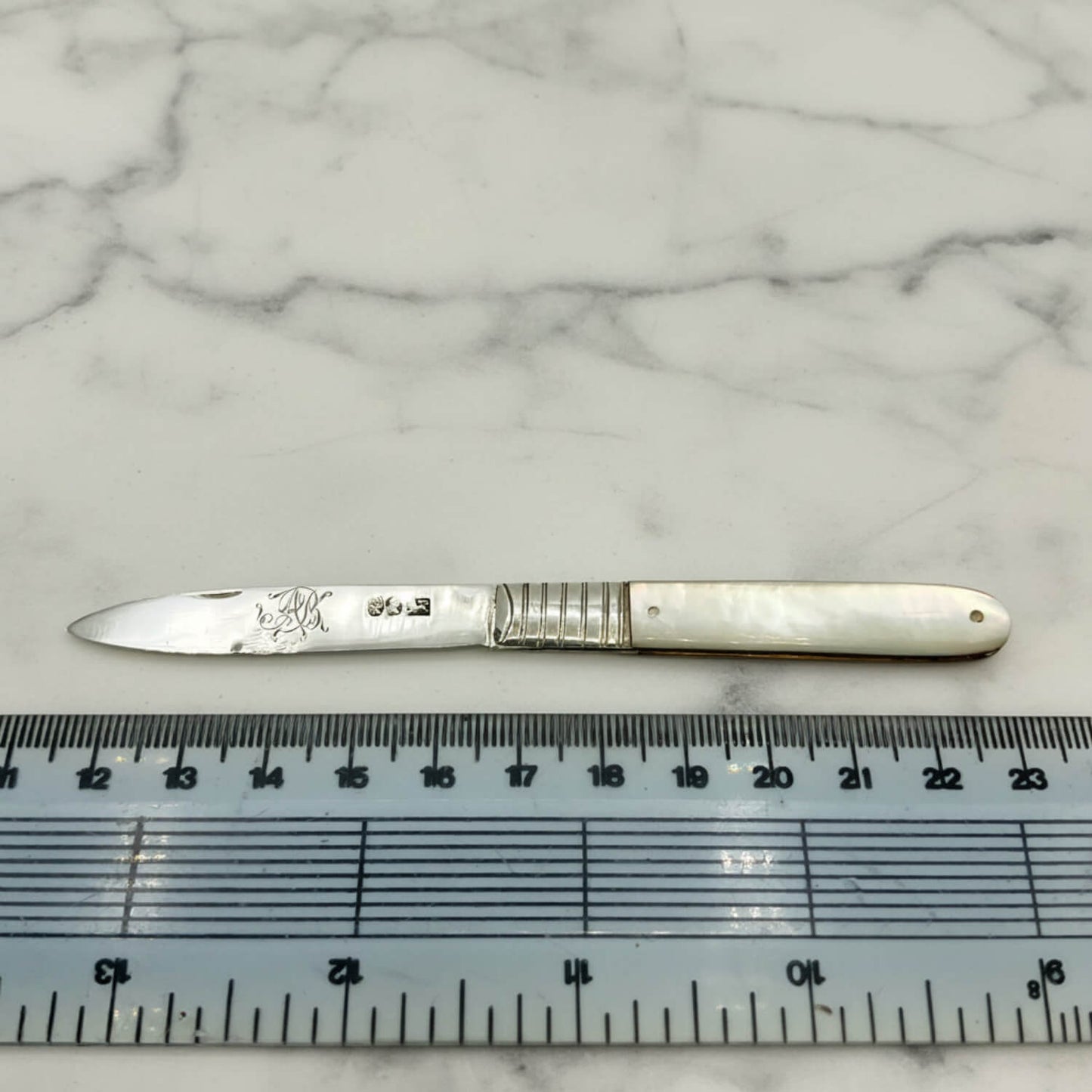 Antique fruit knife with a mother-of-pearl handle, placed horizontally on a white marble surface with a ruler below it for scale.