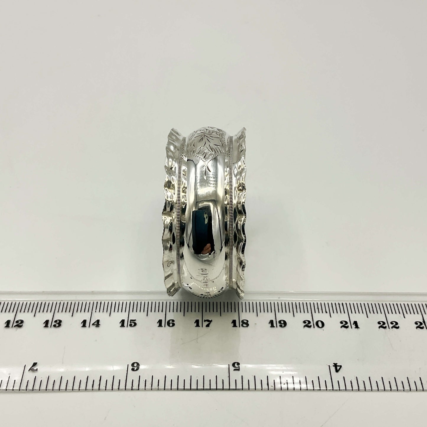Silver napkin ring with intricate design on a white background with a ruler for scale.