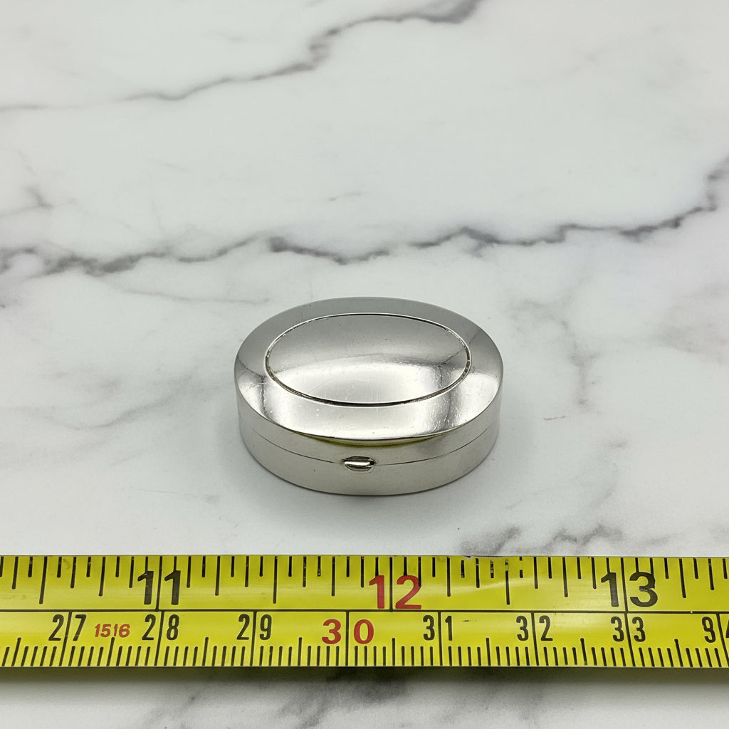 Silver pill box on marble surface next to tape measure for scale