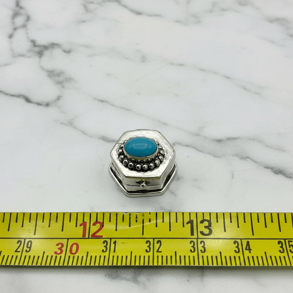 silver pill box next to tape measure for scale on a marble surface