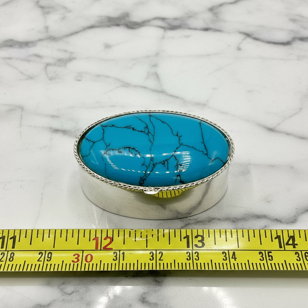 Turquoise stone in a silver pill box with a measuring tape for scale on a marble background