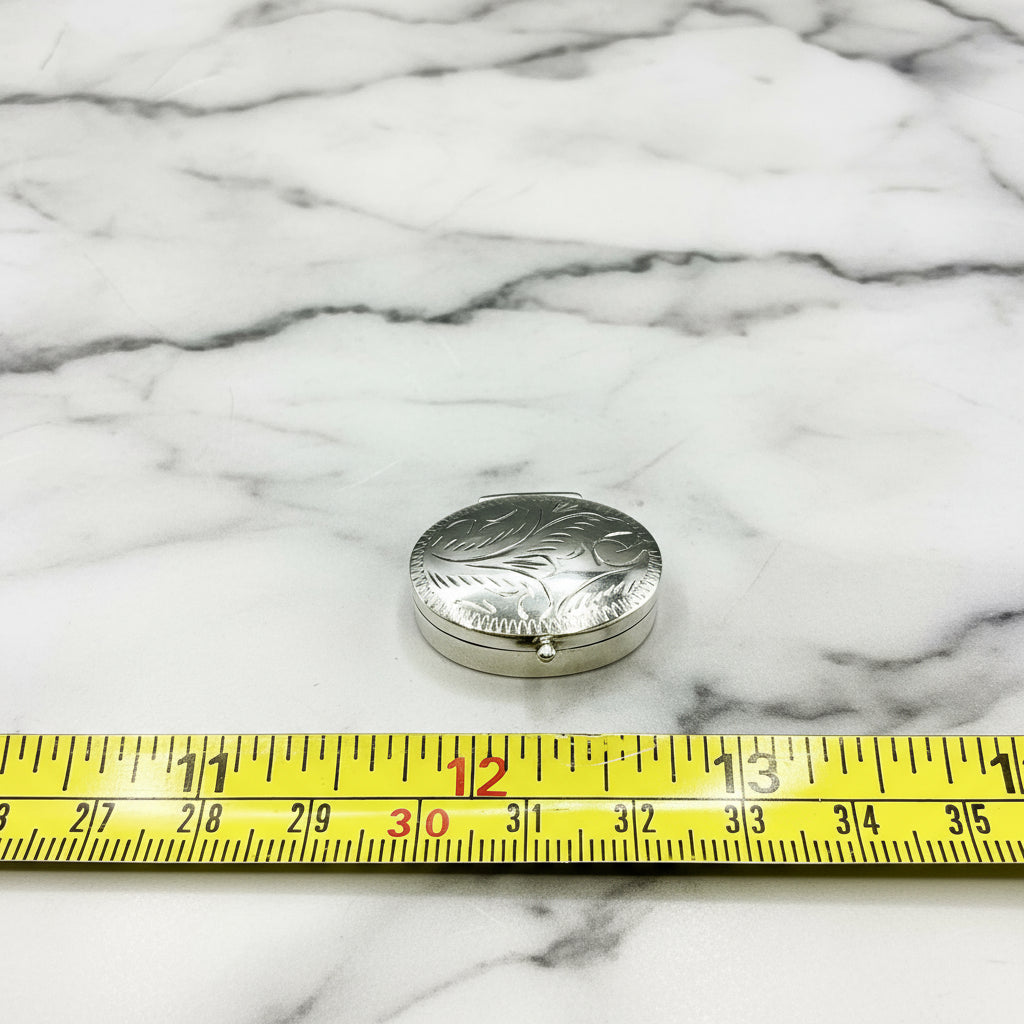 Silver pill box with engraved design on a marble background, measured by a yellow ruler.