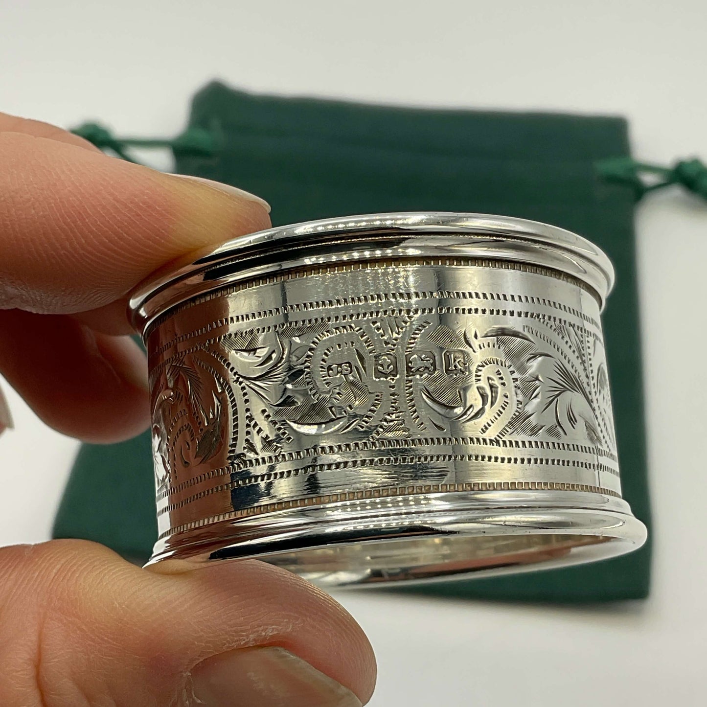 The maker’s mark and hallmarks on the back of an Antique silver napkin ring held in a hand with a green gift bag and white background
