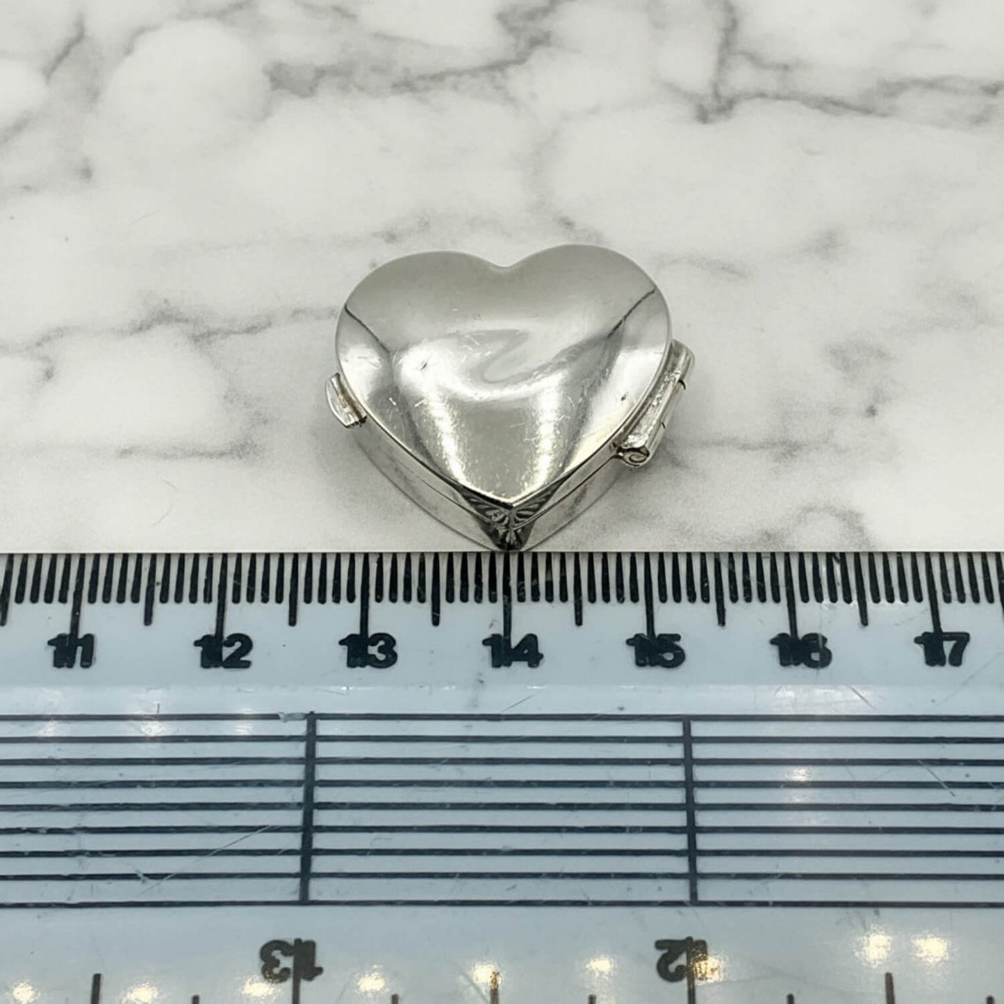Heart-shaped silver pill box on a marble surface with a ruler for scale.
