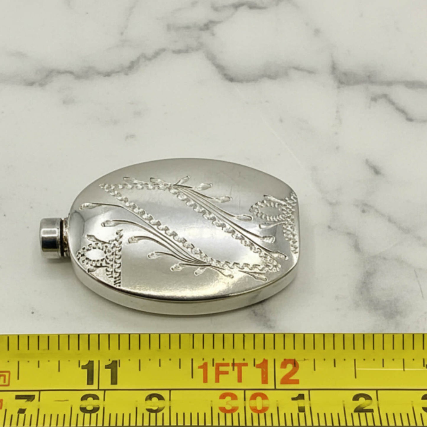 Small Sterling Silver Scent Bottle with engraved design on a marble surface, measured by a yellow ruler.