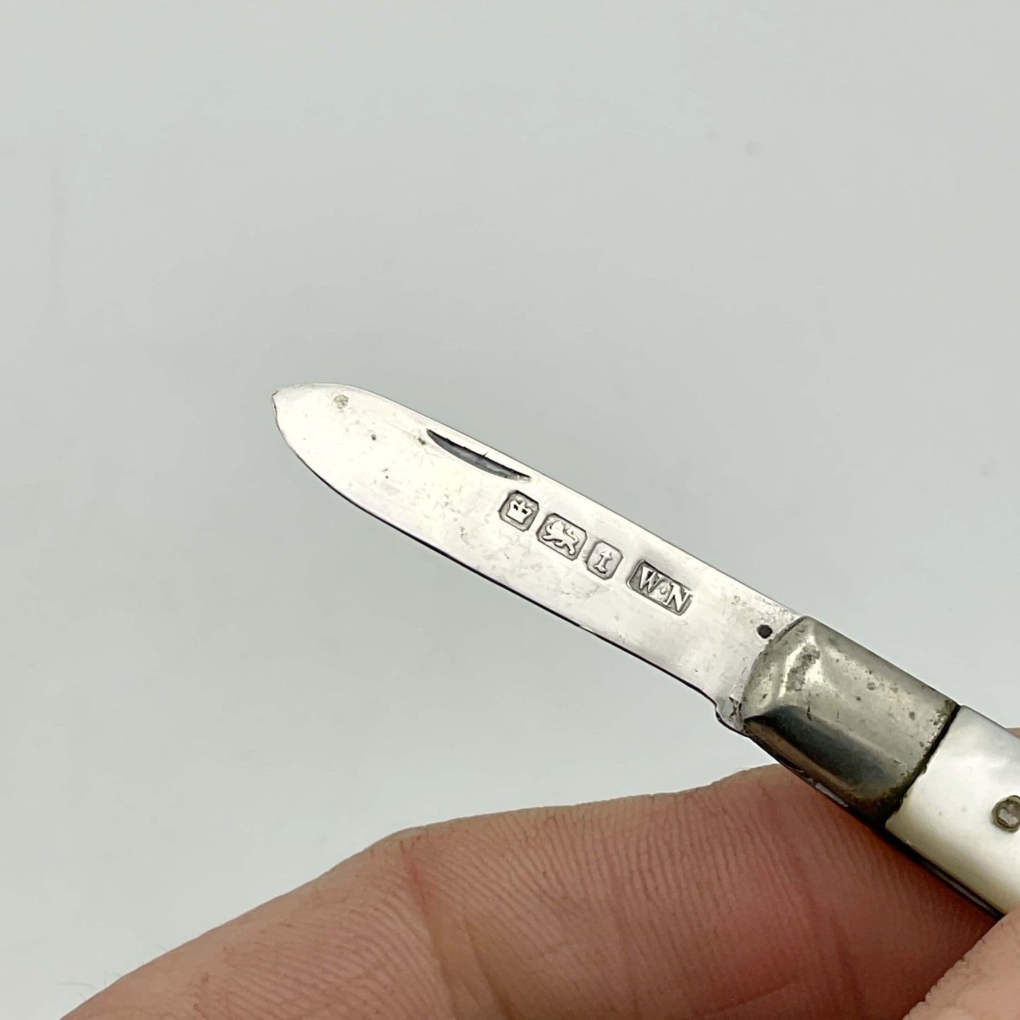 Silver fruit knife with a mother of pearl handle with a close up of the hallmarks on the blade. Held in a hand on a white background.