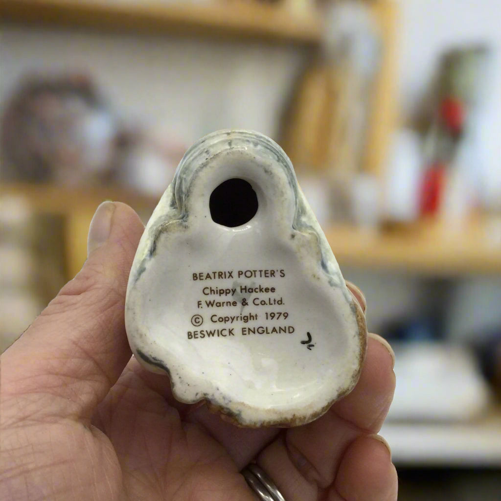 base of figurine with Beatrix Potter and Chippy Hackee on held in a hand with shelving in the background