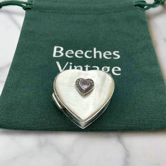 Heart shaped silver pill box with an amethyst and mother of pearl lid on a green bag on a marble background.