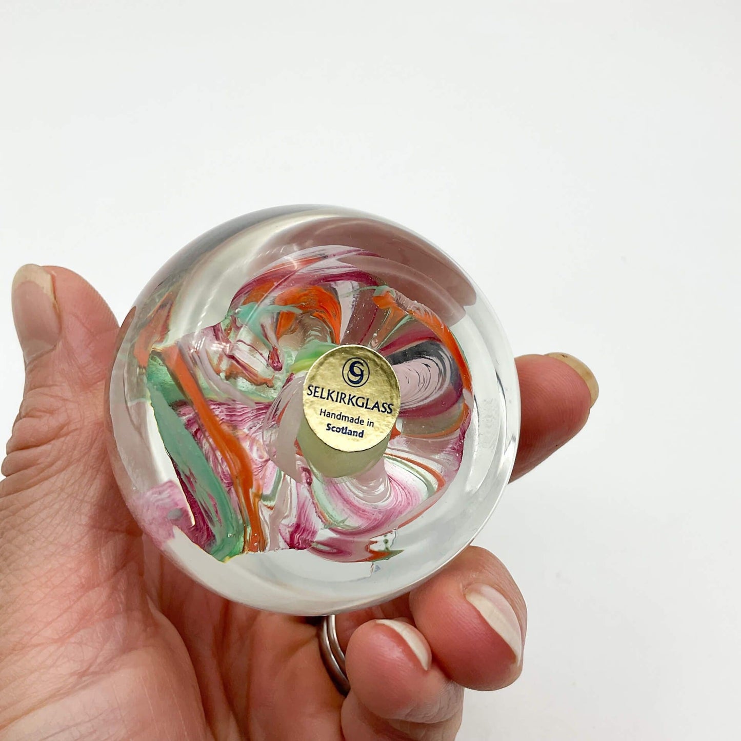 Base of Selkirk glass paperweighrt with multicoloured swirls held of a hand on a white background.