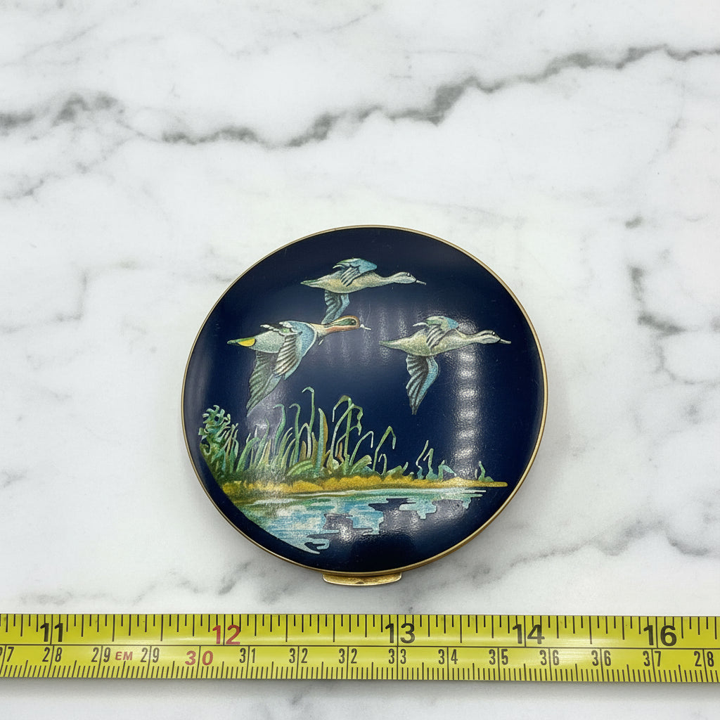 beautiful enamel featuring three ducks flying over water on a mirror compact lid next to a tape measure on a marble surface