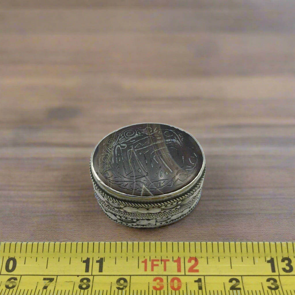 Silver decorative box with intricate designs on a wooden surface, measured by a yellow tape measure.