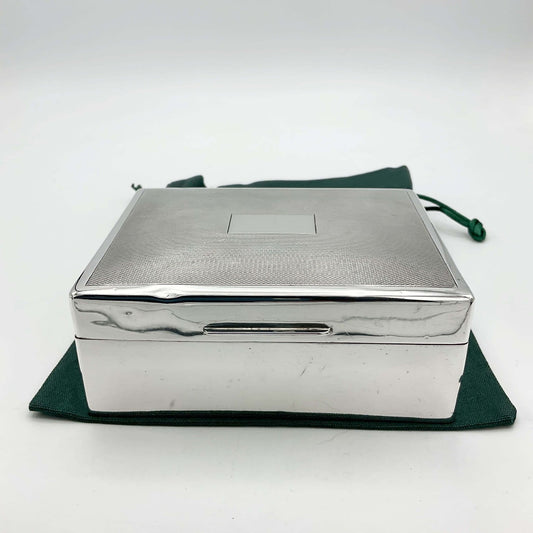 Vintage Sterling Silver cigarette box with shiny silver sides and a patterned lid sitting on a green cotton bag. The lid has a blank rectangular square in the centre.