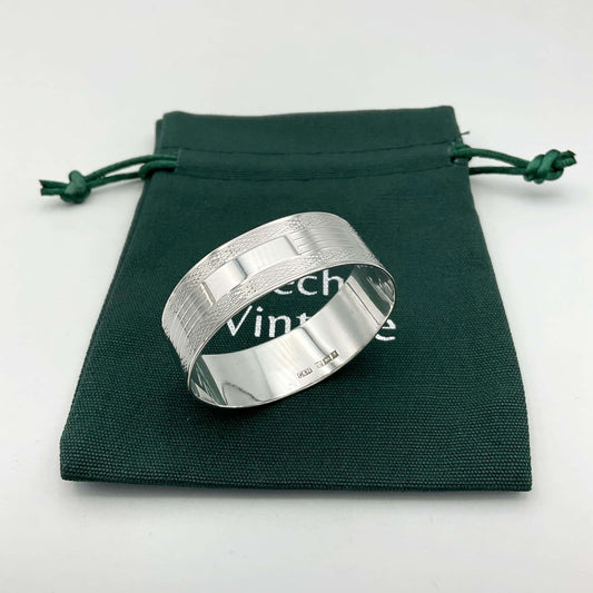 Shiny silver napkin ring with a lined pattern around the sides and a blank rectangle in the centre. It is sitting on a green Beeches Vintage cotton bag.