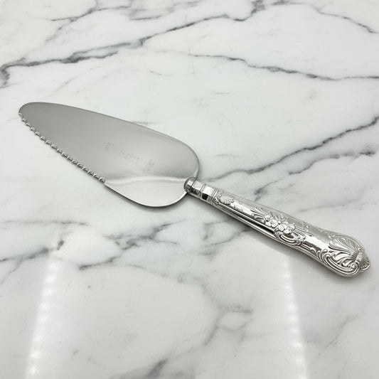 A silver coloured metal cake or pie slice on a marble background. The handle has a Queens pattern elaborate design.