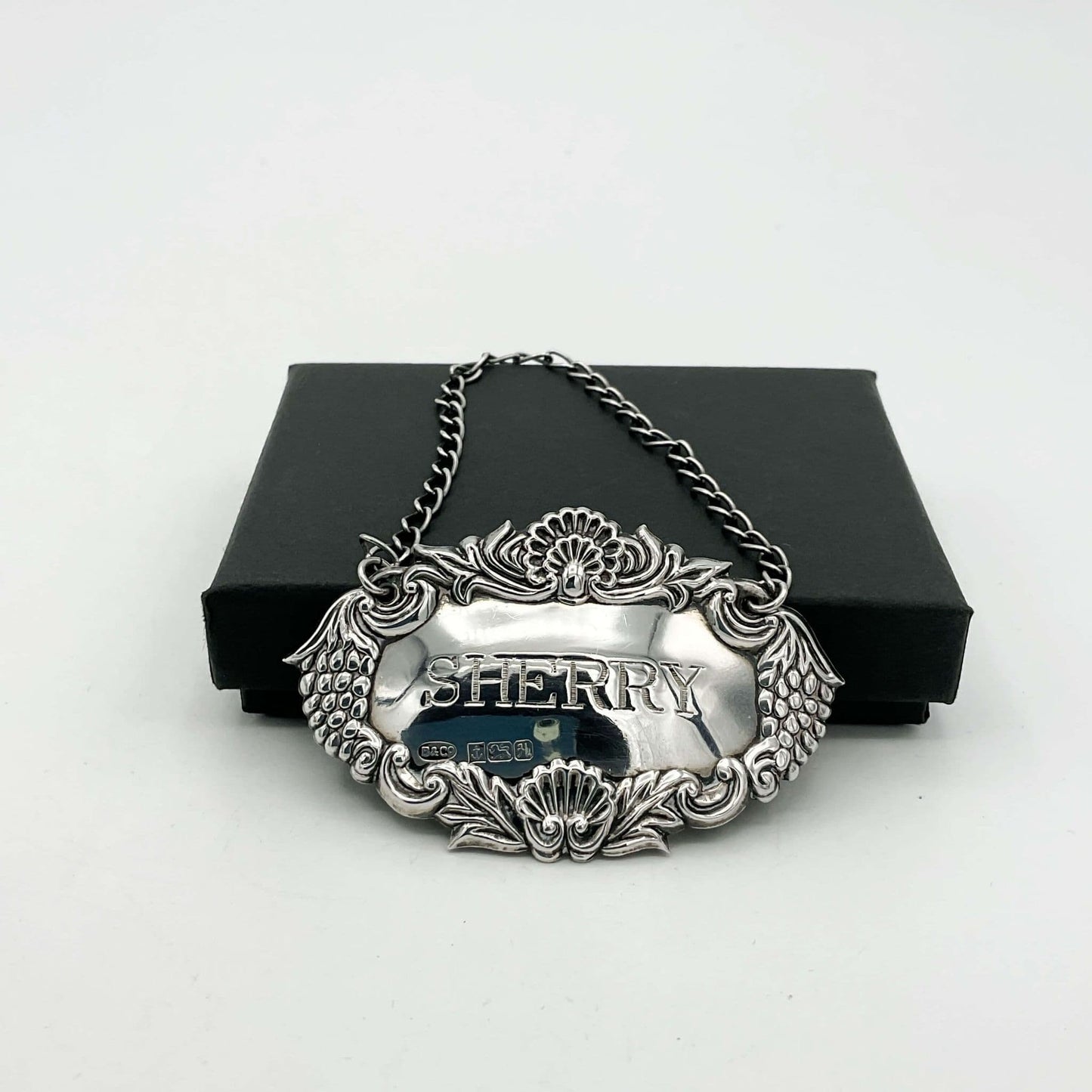 Silver decanter lable with a grapes pattern on sides and Sherry in the middle leaning against a black box