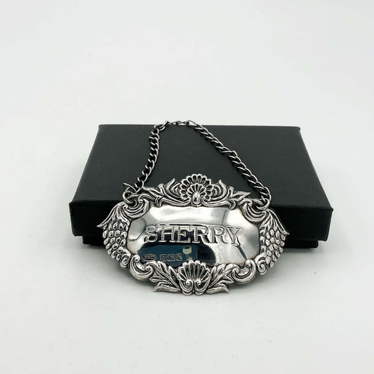 Silver decanter lable with a grapes pattern on sides and Sherry in the middle leaning against a black box