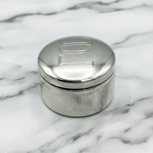 A polished sterling silver pill box with the Vourakis brand mark stamped on the base, featuring Greek letters engraved on the lid on marble surface
