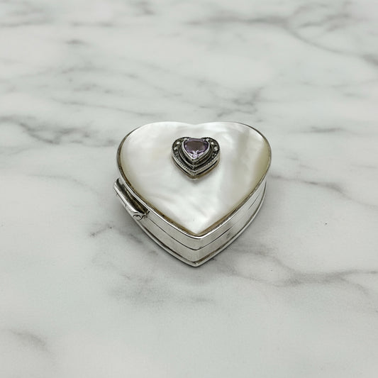 Heart shaped silver pill box with an amethyst and mother of pearl lid on a marble background.