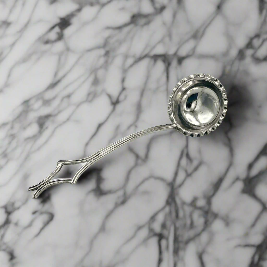 A silver-plated sauce ladle with a crimped pattern edge bowl and a diamond pattern handle on a marble surface.