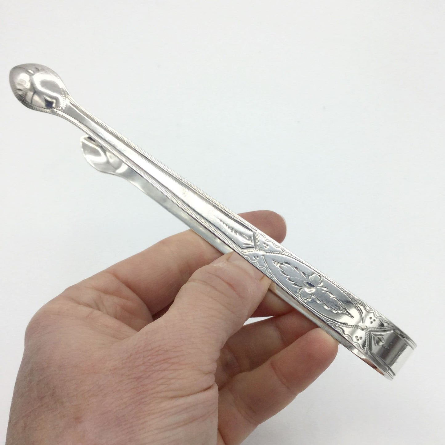 1700s Silver sugar tongs with a pretty floral engraved design on a white background held in hand
