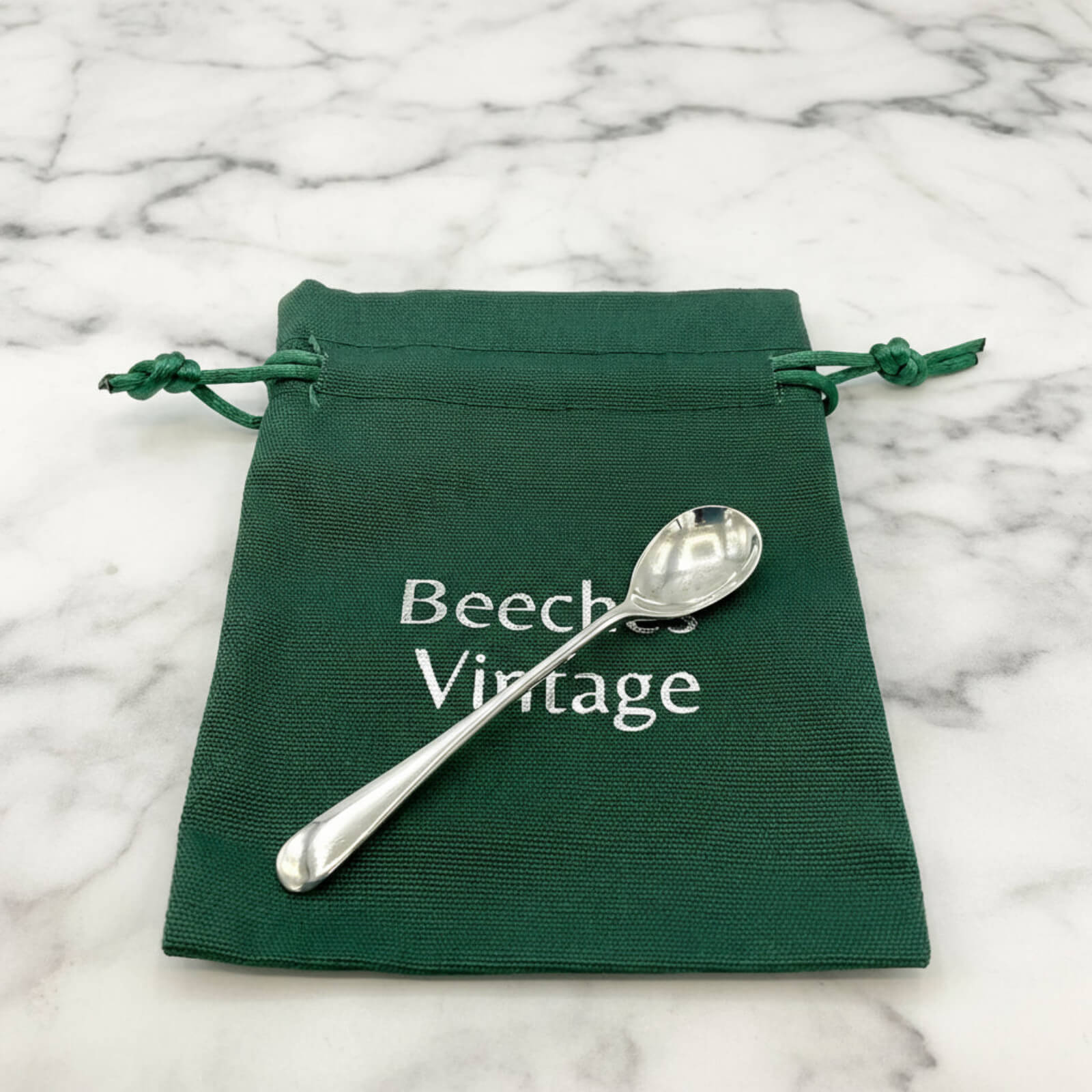 Silver spoon on a green drawstring bag with 'Beeches Vintage' branding on a marble background.