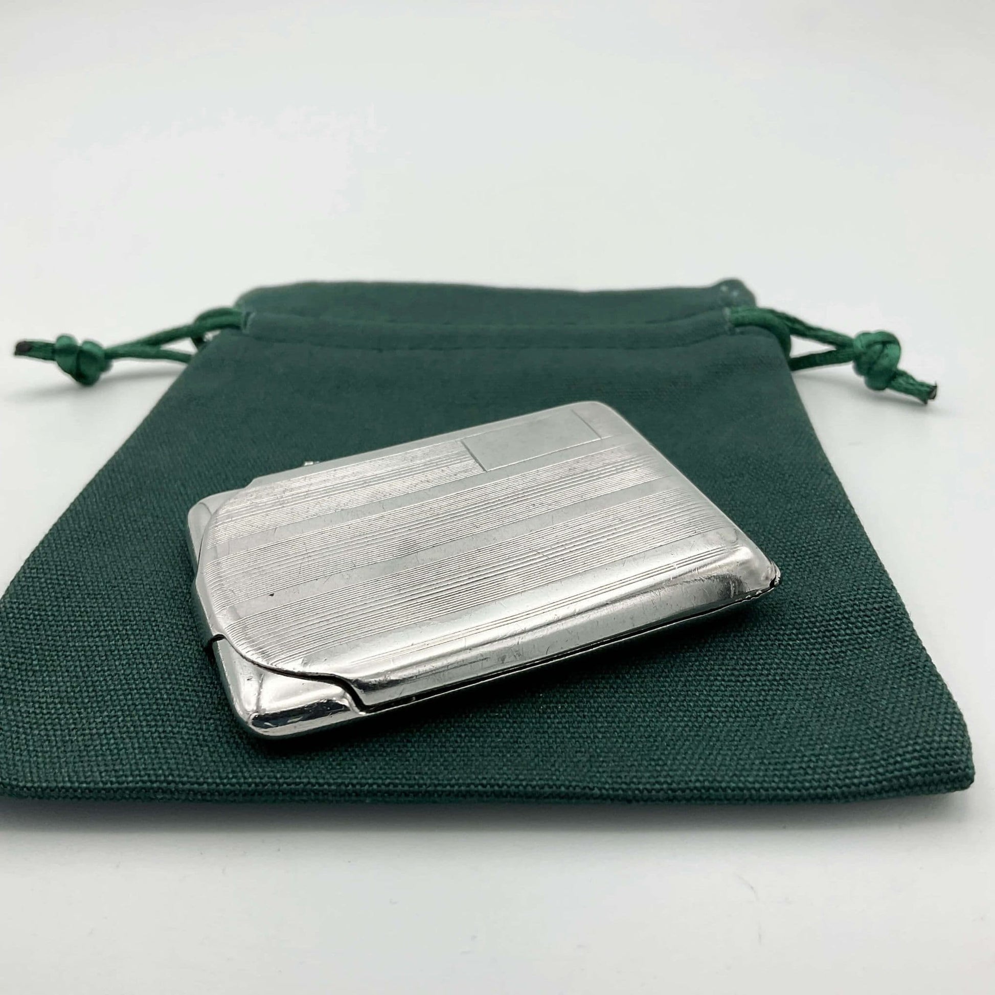 Side view of Silver matchbook case with a lined pattern and blank box for engraving initials in left hand corner on a green gift bag.