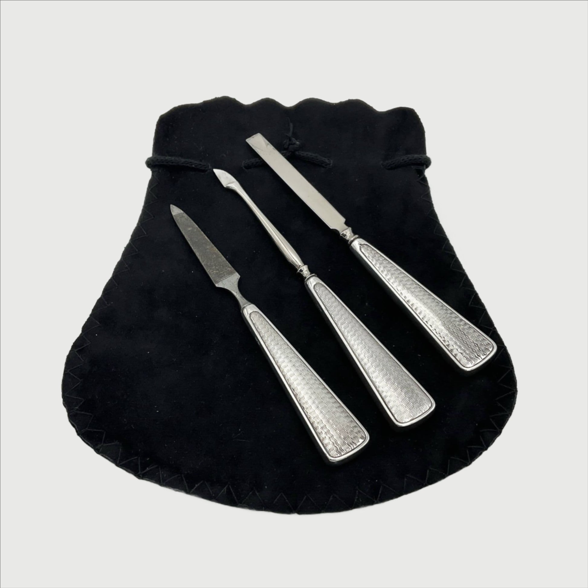 Three manicure tools with wavy 1930s design on the silver handles. The tools are a nail file and two types of cuticle pushers on a black bag.