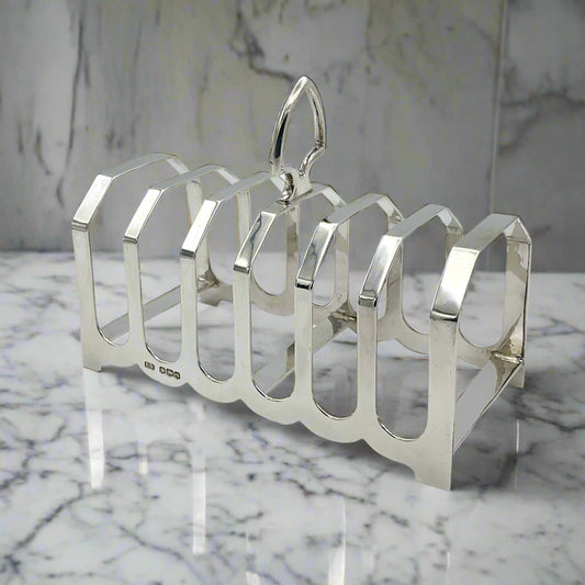 A vintage Art Deco sterling silver toast rack with a loop handle on the middle arch and seven arches, displayed against a marble background.