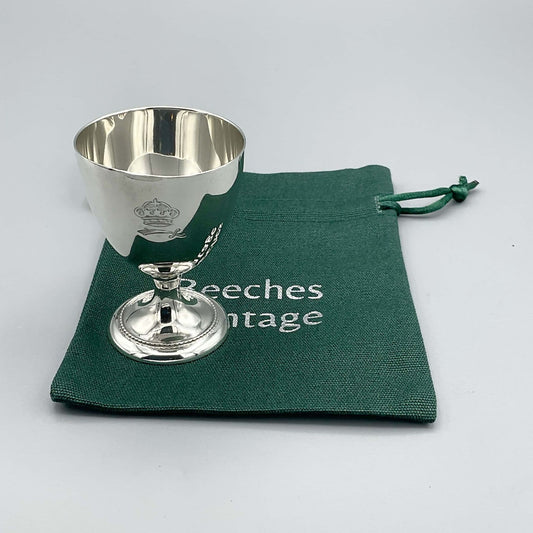 Silver pedestal shaped egg cup standing on a green Beeches Vintage cotton bag. The side of the egg cup shows a crown and sword motif.
