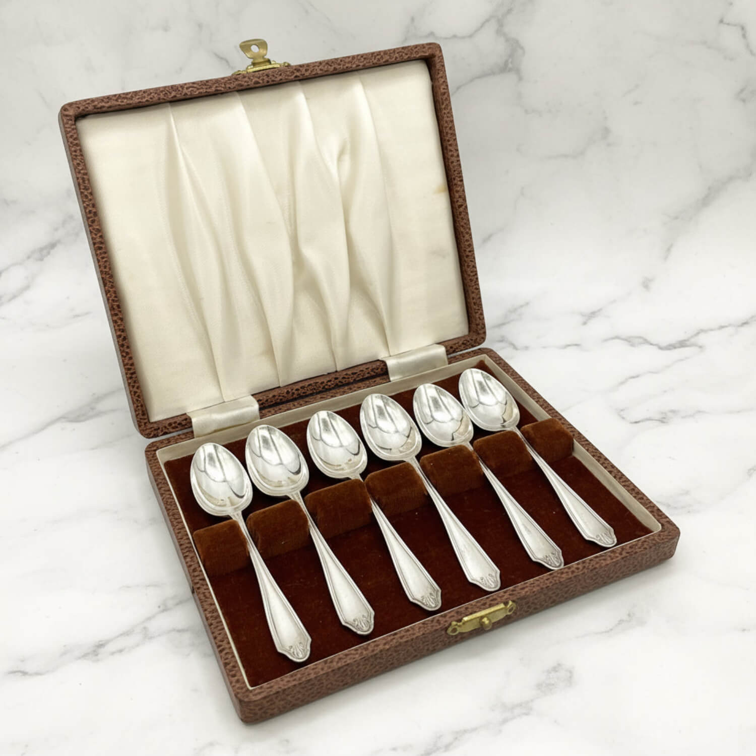 An open brown textured presentation box containing six silver coffee spoons on a white marble surface. The box has cream-colored fabric lining on the inside lid and a dark brown velvet-like base holding the spoons.