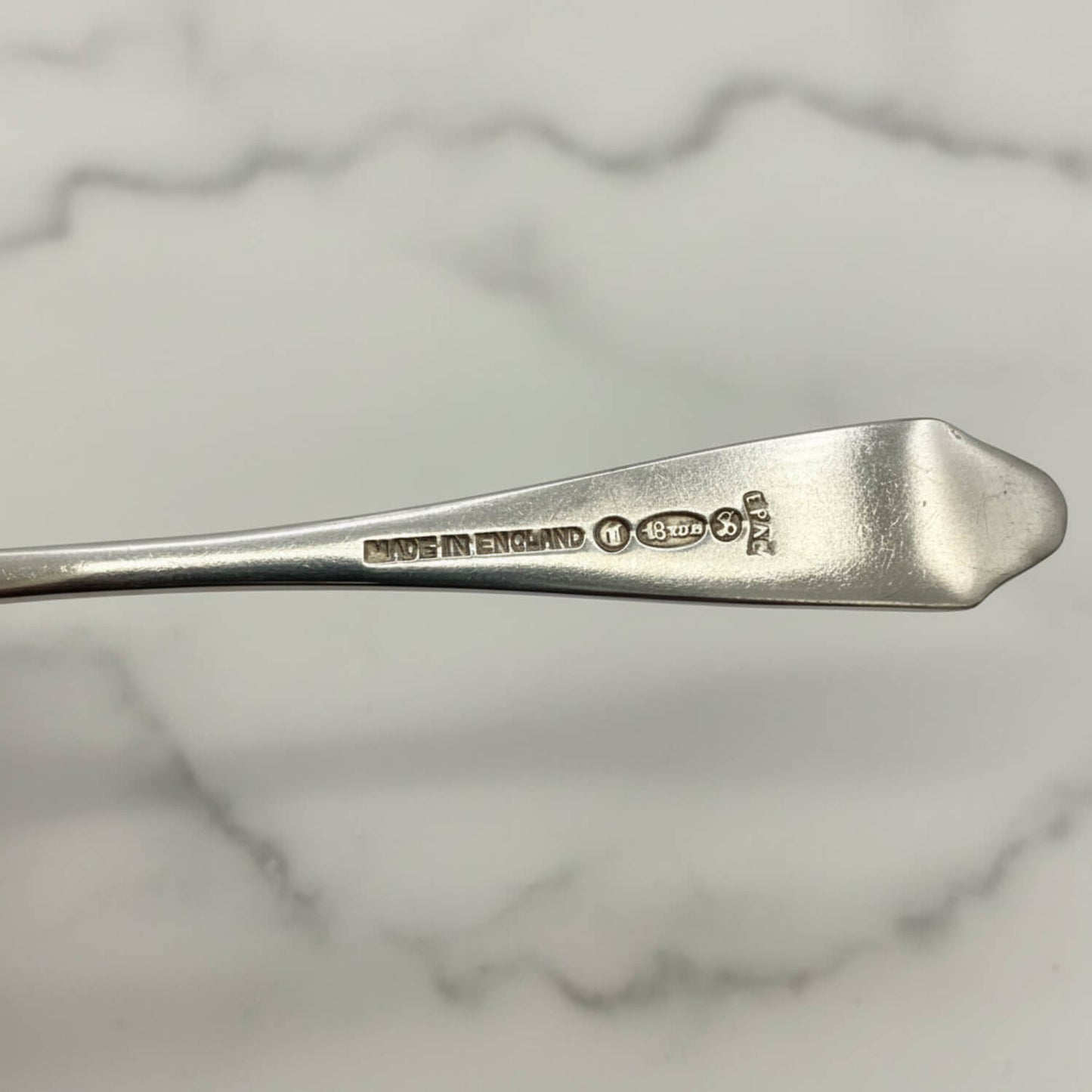 A close-up of a silver coffee spoon handle with engraved hallmarks including 'MADE IN ENGLAND' and makers marks, set against a white marble background.