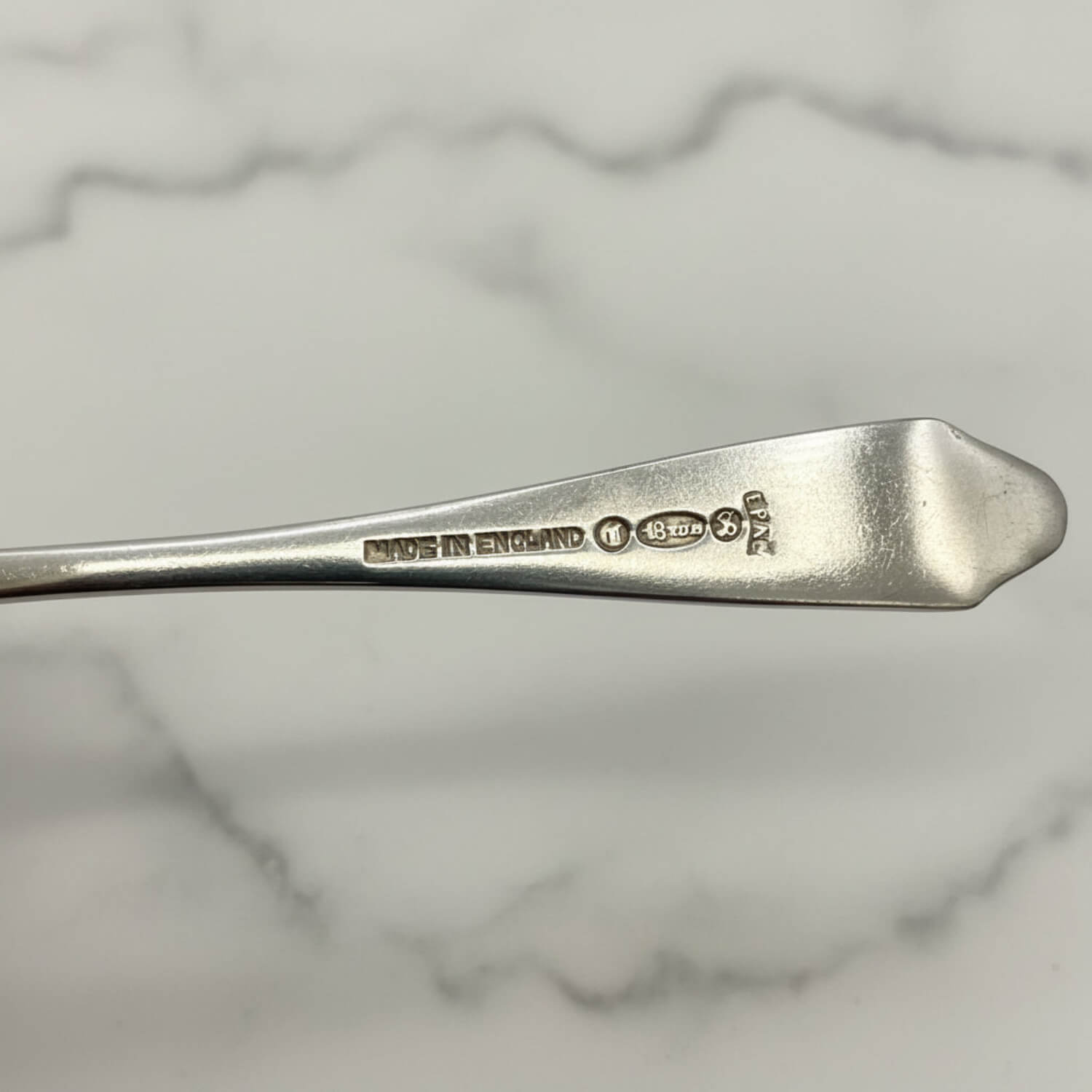 A close-up of a silver coffee spoon handle with engraved hallmarks including 'MADE IN ENGLAND' and makers marks, set against a white marble background.