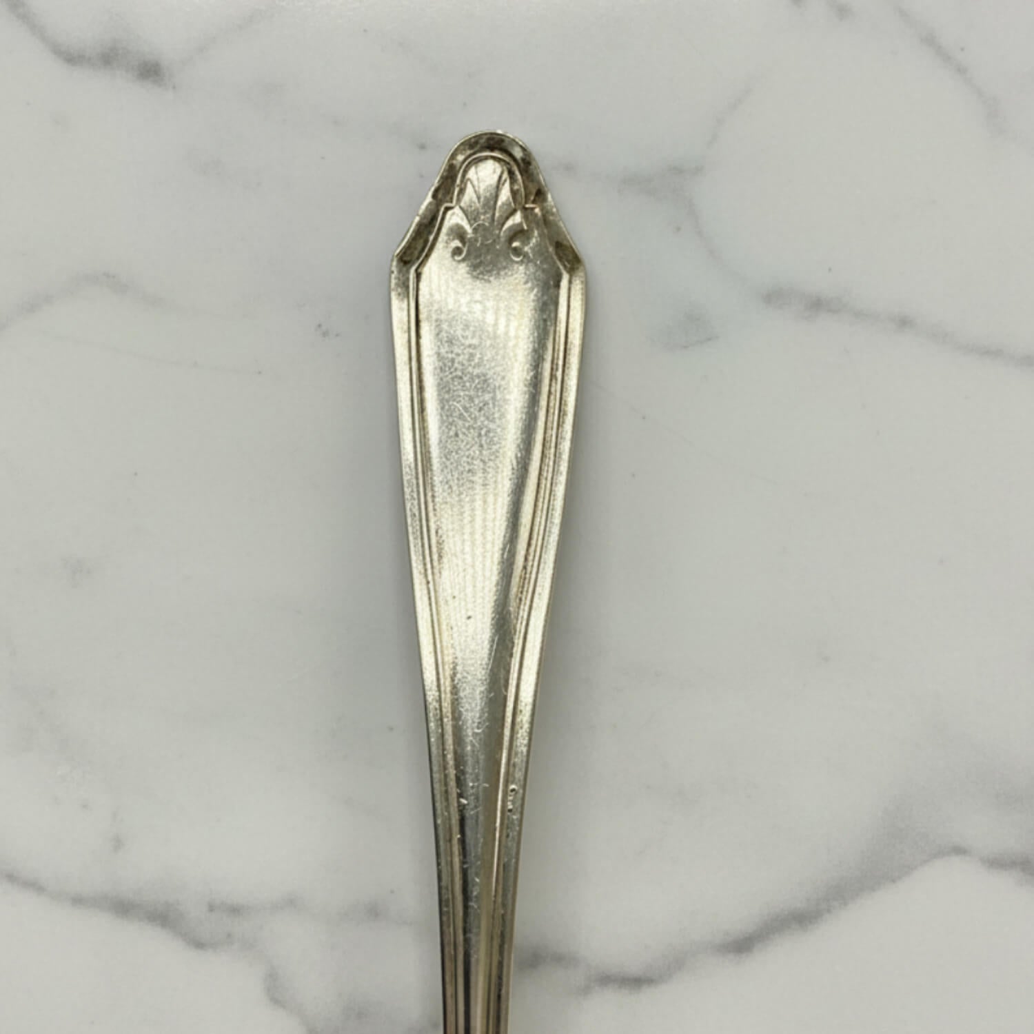 A close-up of a polished silver coffee spoon handle with a decorative crest at the top, resting on a white marble surface with gray veining.