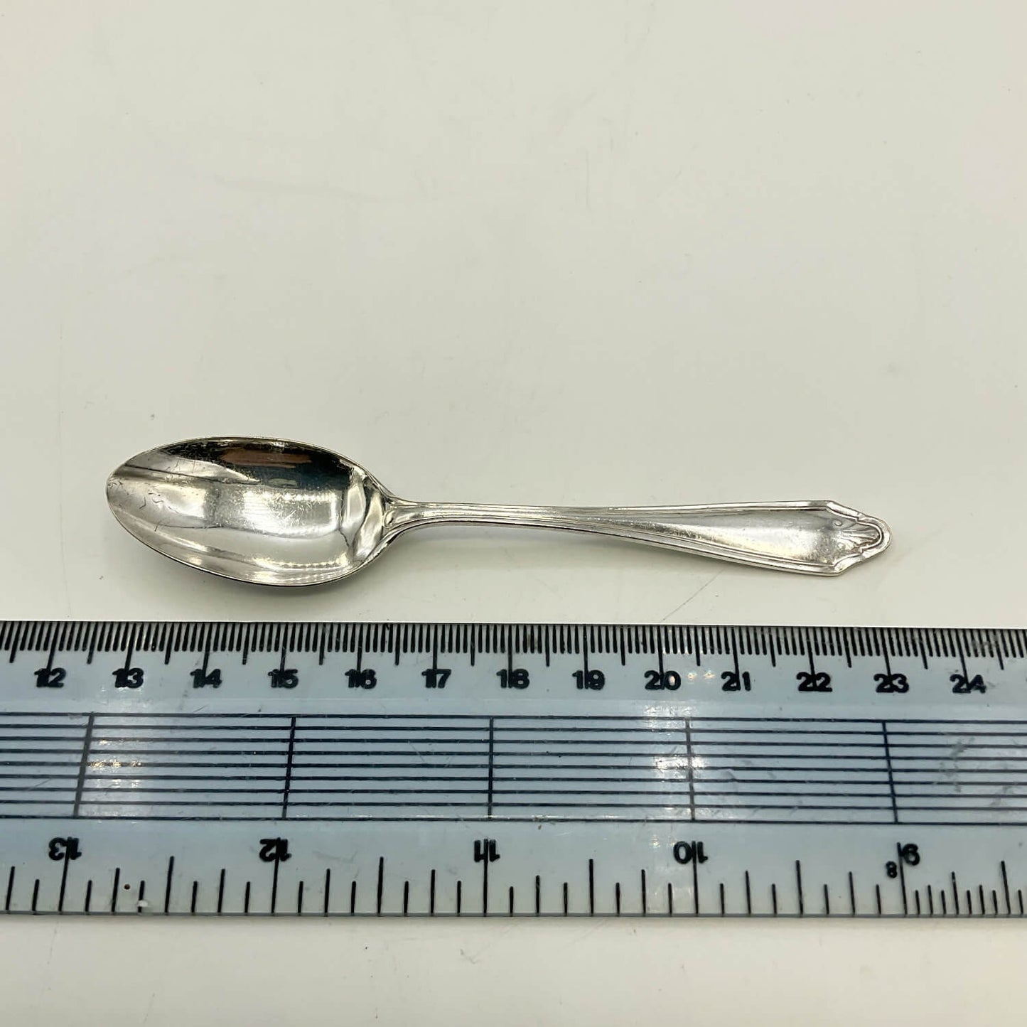 A vintage silver plated coffee spoon with a decorative handle, shown horizontally above a clear plastic ruler for scale on a white background.