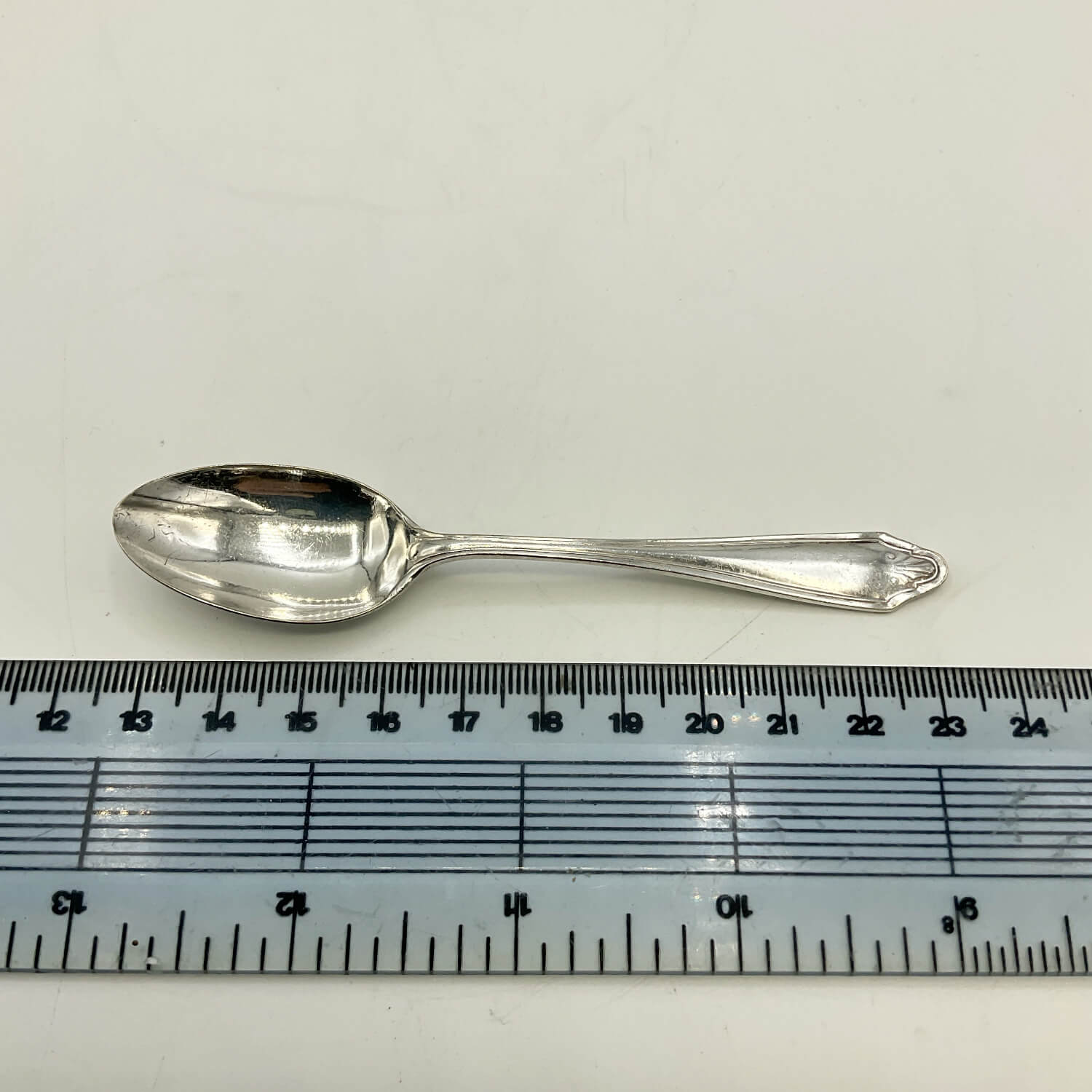 A vintage silver plated coffee spoon with a decorative handle, shown horizontally above a clear plastic ruler for scale on a white background.