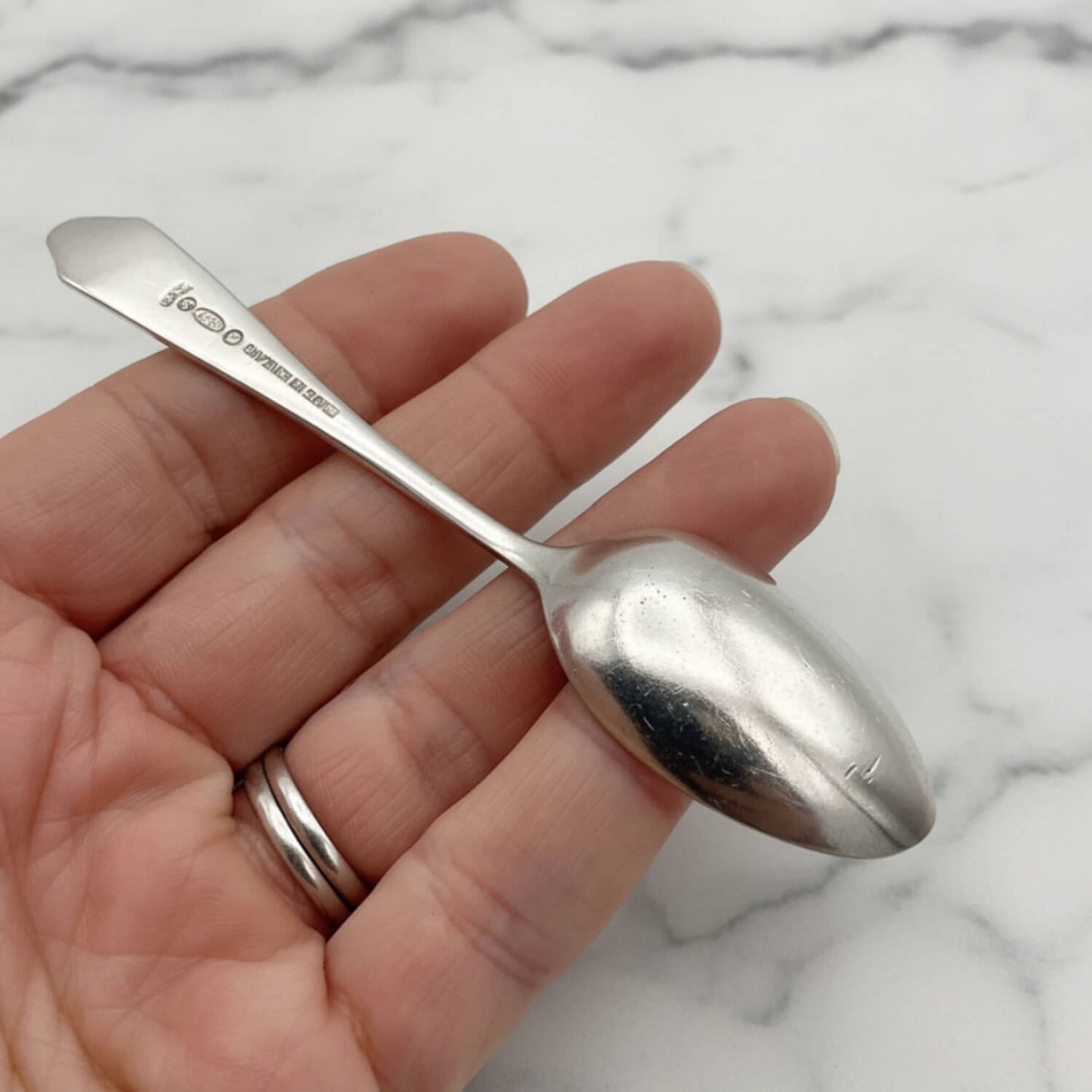 A small silver coffee spoon held in a person's hand against a white marble background.