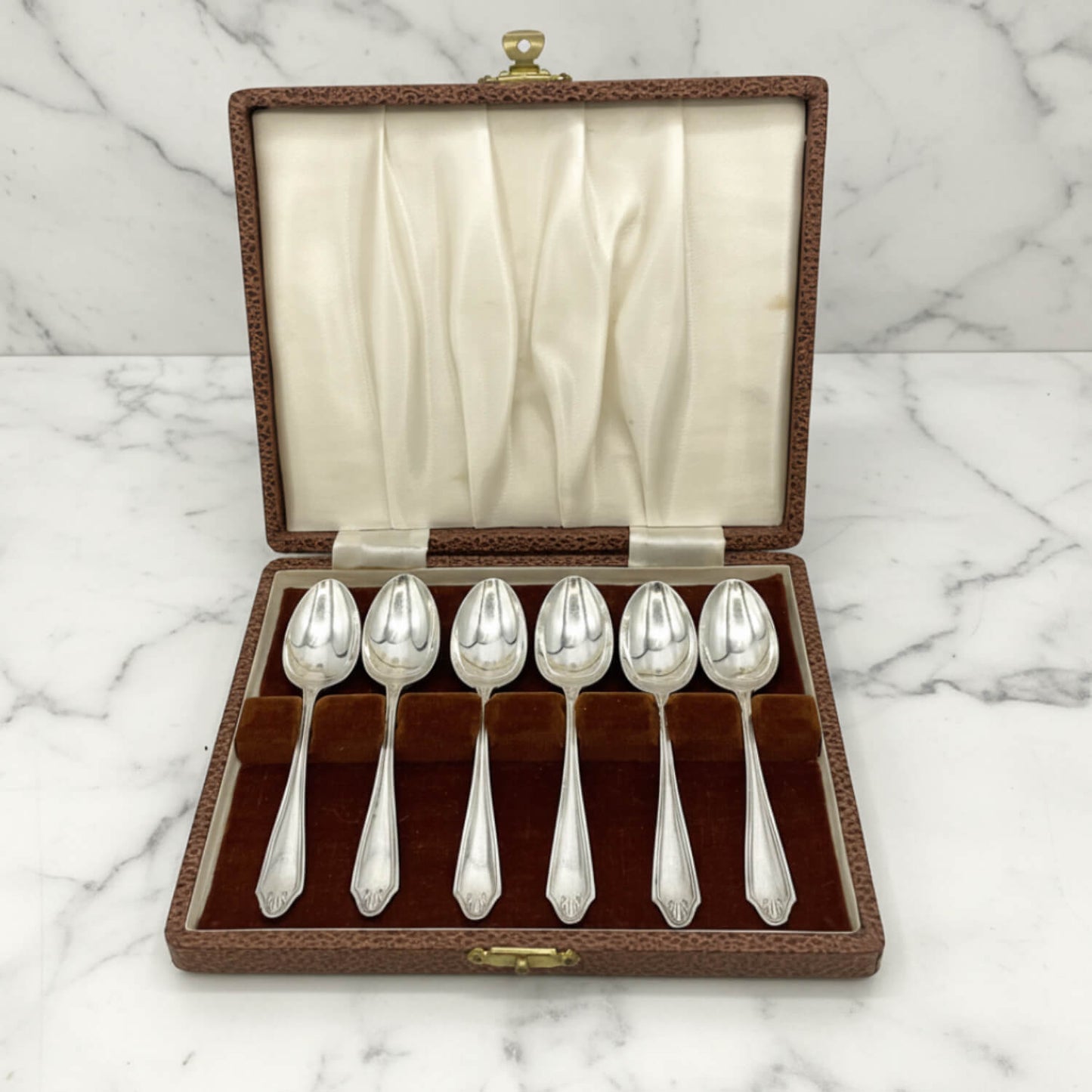 Six polished silver coffee spoons displayed in a brown textured presentation box with a cream satin-lined lid, set on a white marble surface.