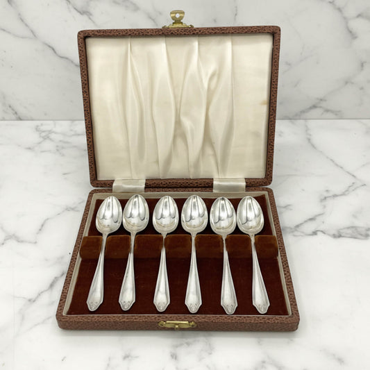 Six polished silver coffee spoons displayed in a brown textured presentation box with a cream satin-lined lid, set on a white marble surface.