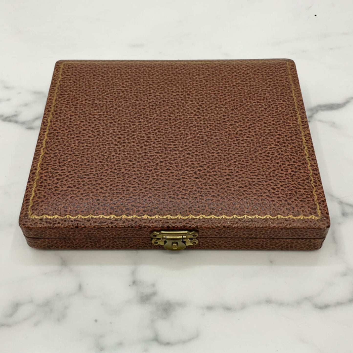 A rectangular brown textured leather case with gold-colored stitching and a brass clasp, resting on a white marble surface.