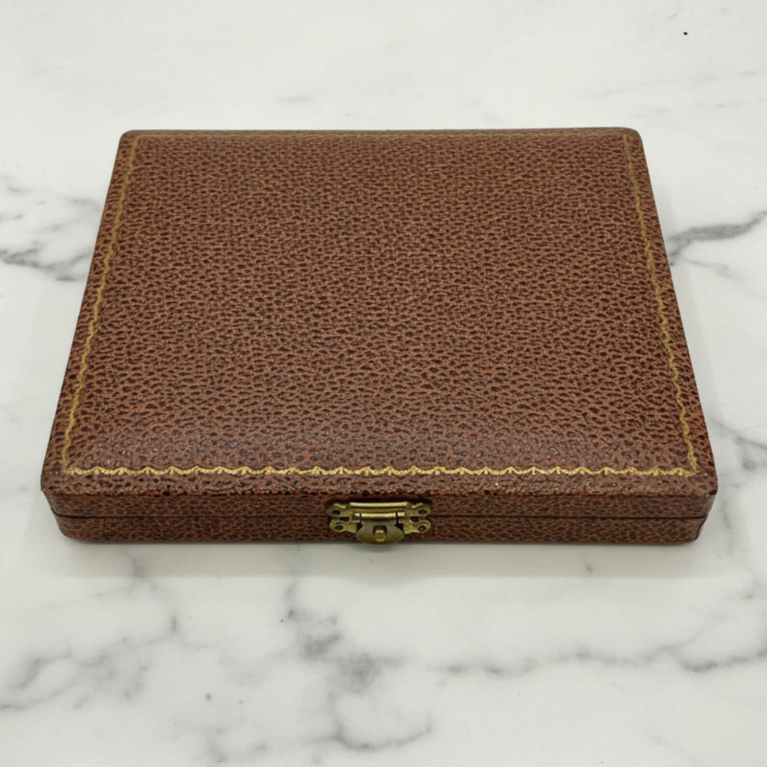 A rectangular brown textured leather case with gold-colored stitching and a brass clasp, resting on a white marble surface.