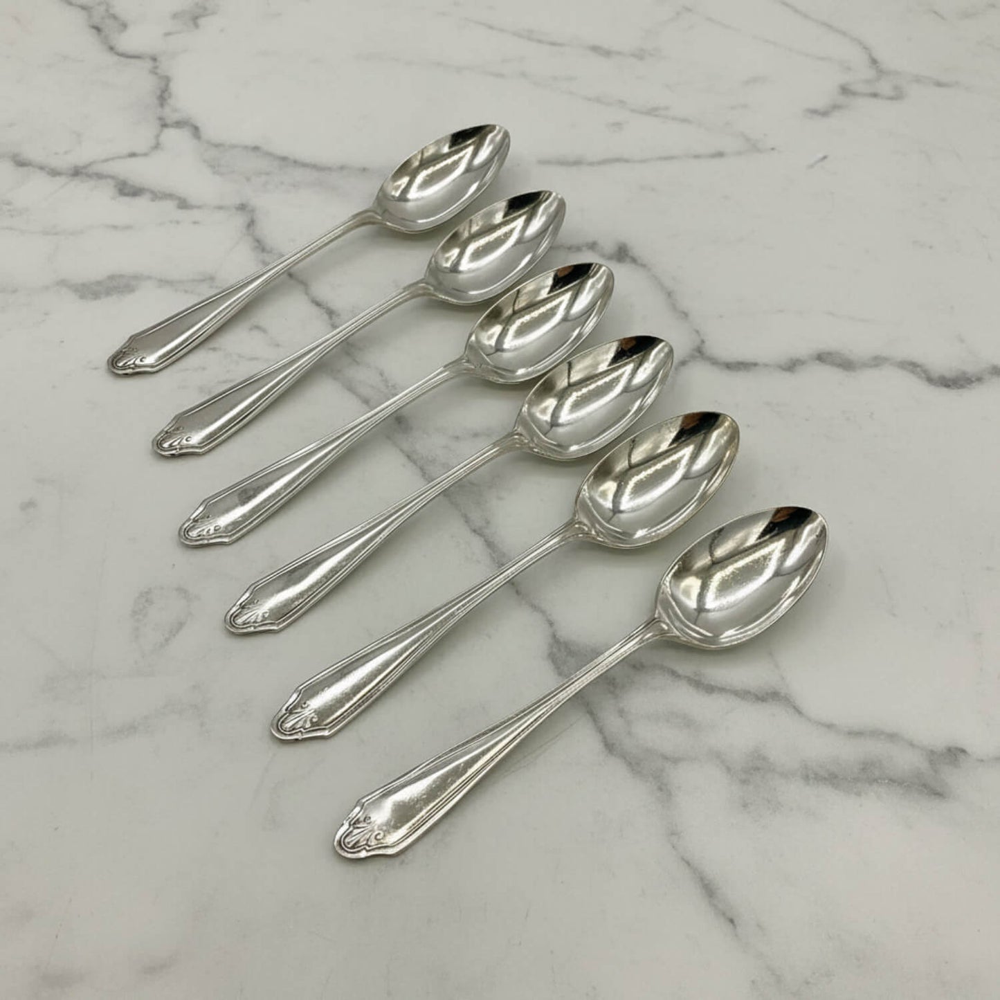 Six polished silver coffee spoons arranged diagonally on a white marble surface with gray veining.