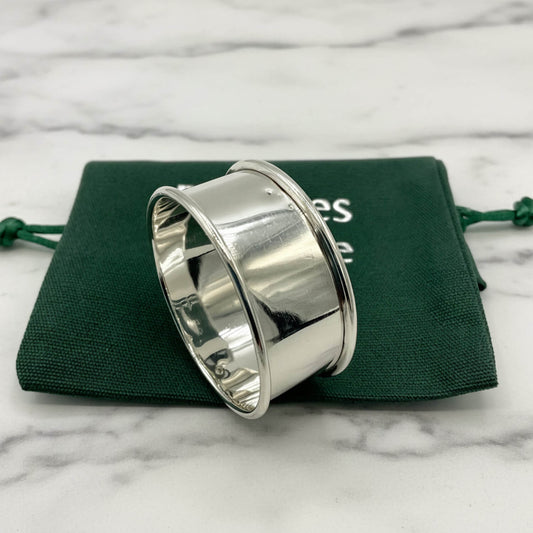 A polished silver napkin ring with a wide, flared band, displayed on top of a dark green drawstring pouch against a white background.