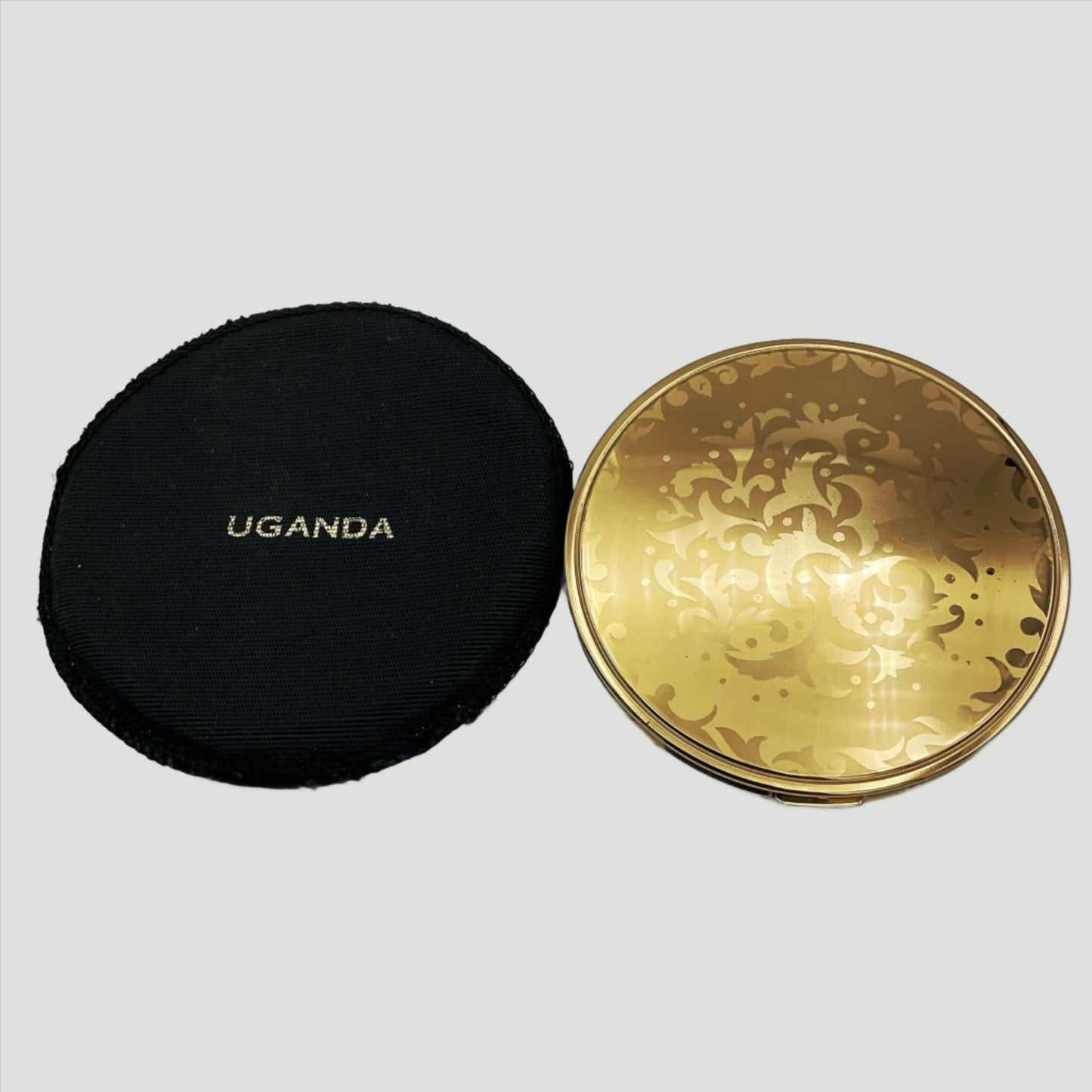 gold coloured lid of powder compact with leaf pattern on a black felt case with Uganda imprinted on it.