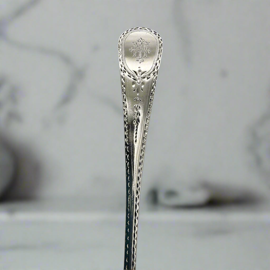 Silver serving spoon handle showing bright cut design and engraved initials at top with a marble background