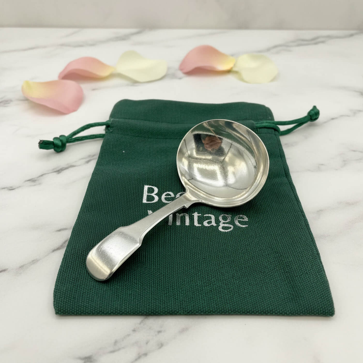 Antique 1800s Silver Tea Caddy Spoon on a green bag on a marble surface with petals in the background