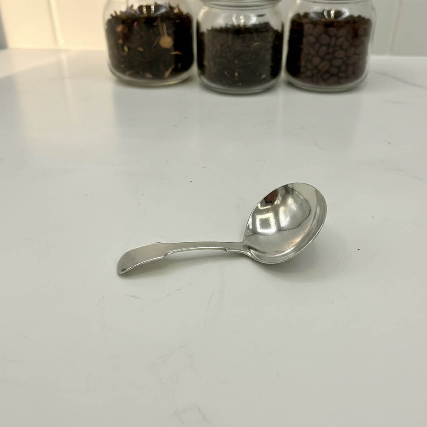Antique 1800s Silver Tea Caddy Spoon on a marble worktop with tea and coffee jars in the background