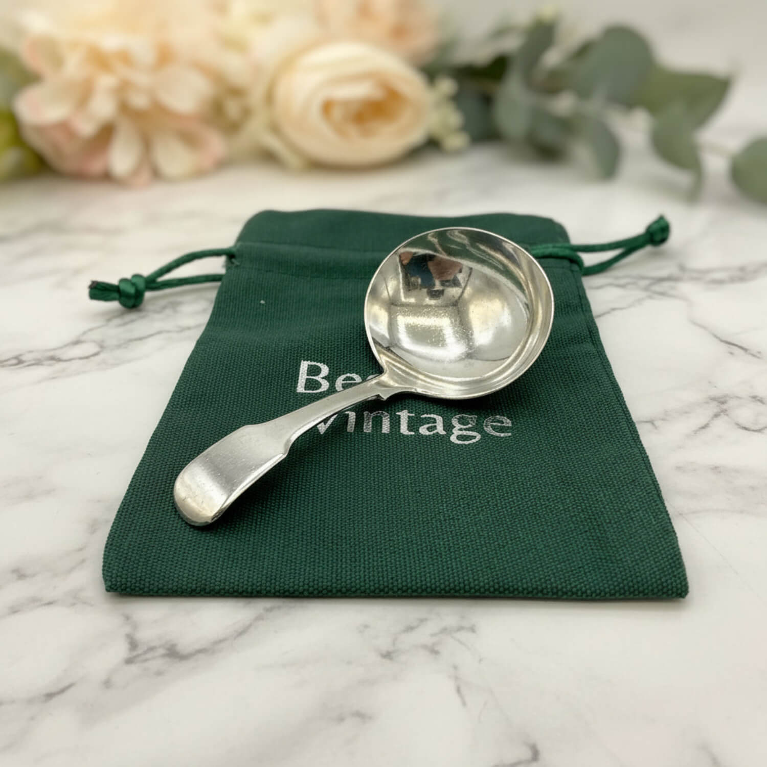 Antique 1800s Silver Tea Caddy Spoon on a green bag on a marble worktop with roses in the background
