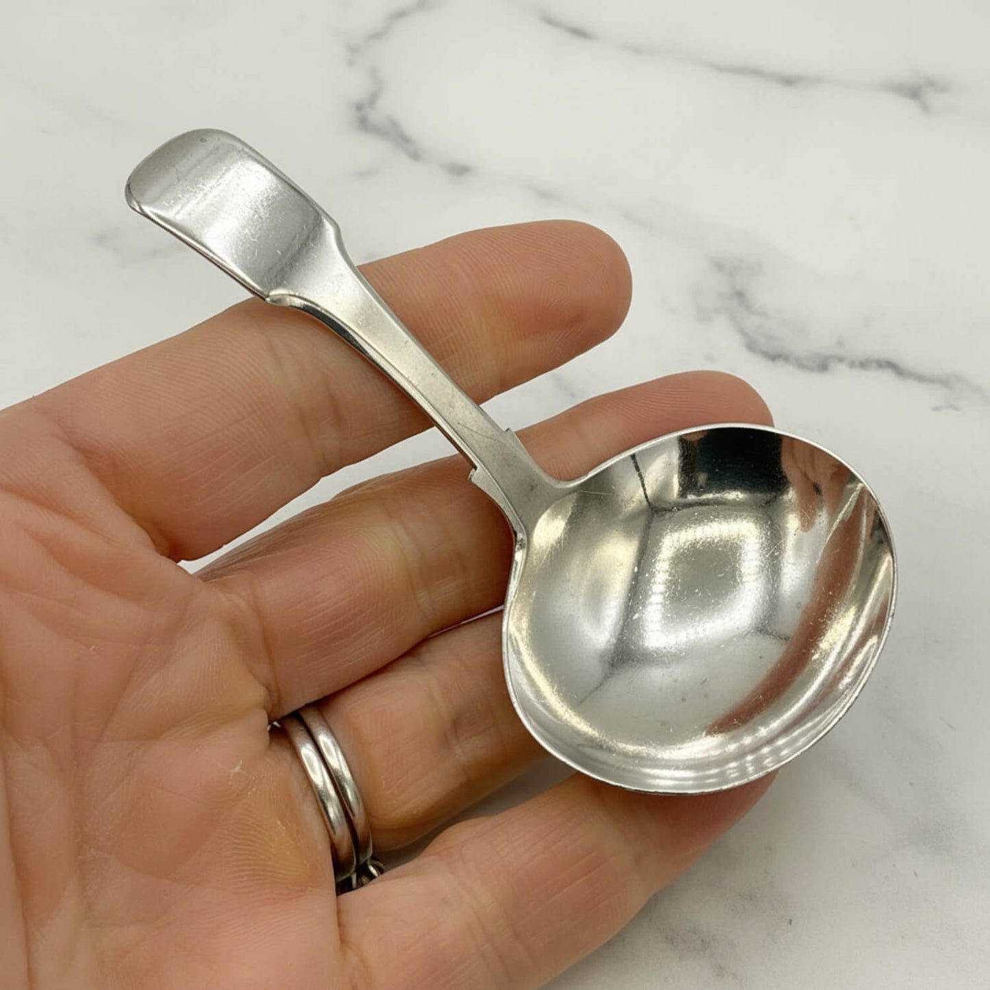 Antique 1800s Silver Tea Caddy Spoon held in a hand with a marble background