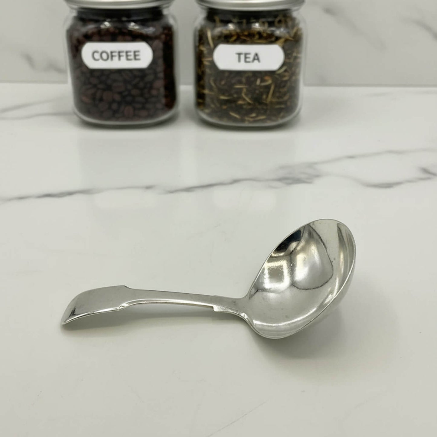 Silver tea caddy spoon on a marble surface with jars labeled 'Coffee' and 'Tea' in the background.